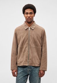 Barna velúr dzseki gallérral, elülső cipzárral és minimális varrással. Kék farmernadrággal párosítva, rétegezett megjelenésért.