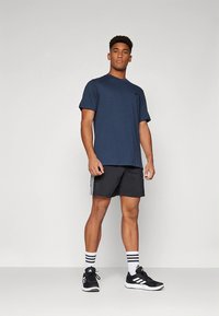 Mörkblå t-shirt med korta ärmar kombinerad med svarta shorts med tre vita ränder, bärs med svarta sportskor och strumpor med vita ränder.