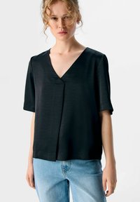 Schwarzes kurzärmliges Oberteil mit V-Ausschnitt, das eine glatte Textur und dezente Faltendetails aufweist. Kombiniert mit hellblauen Jeans.