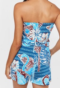Combinaison sans bretelles bleu avec des motifs floraux et de feuillage blancs, mettant en avant des accents rouges. Le tissu a une texture lisse et un design ajusté.