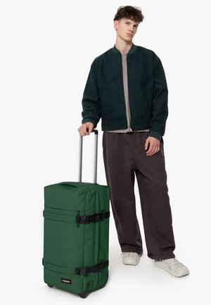 Green soft-shell wheeled luggage with adjustable straps, a zippered compartment, and a sturdy pull handle, positioned beside a person.