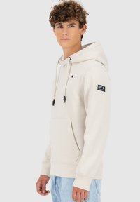 Lichtbeige hoodie van zachte stof met trekkoorden en een zak aan de voorkant. Heeft een zwart logo-patch op de linkermouw.