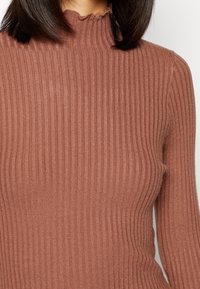 Top a maniche lunghe in rib a terracotta con un collo alto e volant. La texture morbida con righe verticali aggiunge interesse visivo.