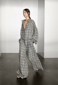 Modelka kráčí sebevědomě v volném, kostkovaném šedém outfitu, který zahrnuje zkrácenou košilku na knoflíky a široké kalhoty na minimalistické scéně.