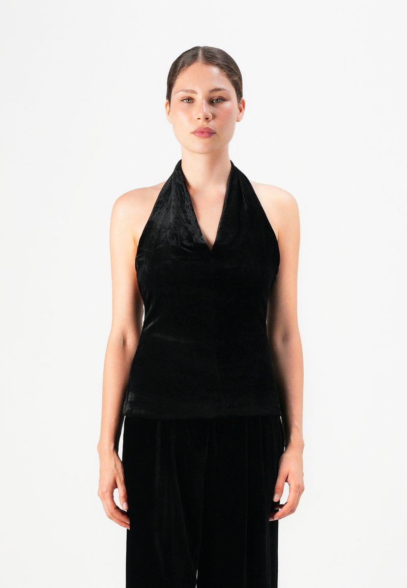Woman standing against a white background wearing a black velvet halter top and matching wide-leg pants.