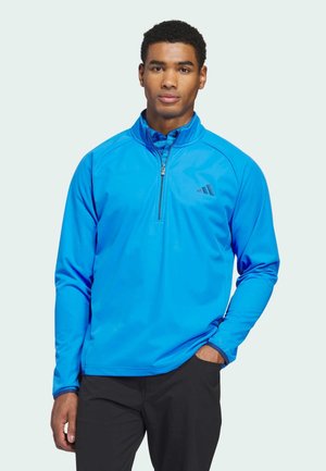 Giovane uomo che indossa un pullover a mezza cerniera di un brillante blu con pantaloni neri, in piedi con una mano in tasca su uno sfondo chiaro uniforme.