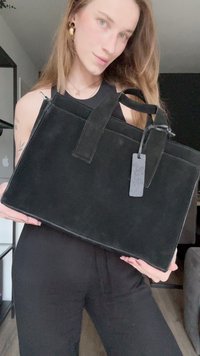 Crna suknena torba s dva gornja ručka, pravokutnog oblika i odvojivom oznakom. Ima glatku teksturu i minimalistički dizajn.