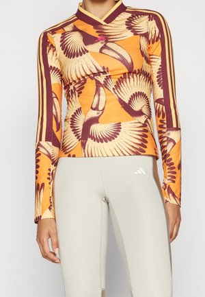 Oranje sporttop met lange mouwen, grote bruine toekan prints en drievoudige bordeauxrode strepen op de mouwen, gecombineerd met lichtbeige legging.
