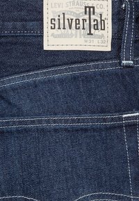 Primer plano de un bolsillo trasero de unos vaqueros de denim azul oscuro con costuras blancas y una etiqueta de Levi Strauss & Co. con el logotipo "silverTab".