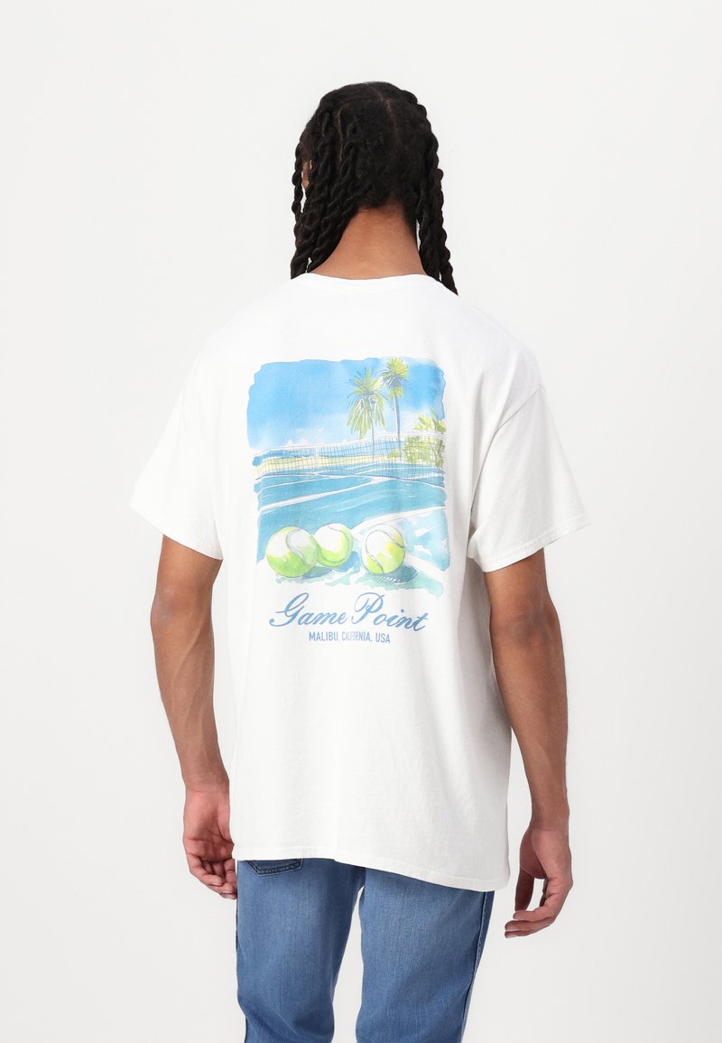 Hvid bomulds t-shirt med et farverigt grafik af tennisbolde og palmetræer mod en blå baggrund, med teksten "Game Point".