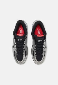 Chaussures de sport noires avec des accents argent métallisé, dotées d'une tige en maille texturée, d'un système de laçage distinctif et de semelles intérieures rouges avec logo.