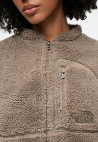 Detailný záber na osobu oblečenú v taupe textúrovanom fleecej mikine s predným zipsom a zipsovaným náprsným vreckom s výšivkou loga The North Face.