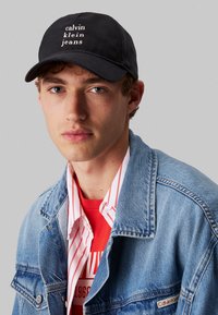 Juodas beisbolo kepurė su baltais "calvin klein jeans" tekstu, dėvima su džinsiniu švarku ant raudonos marškinėlių su baltais dryžiais, pabrėžianti sluoksniavimą.