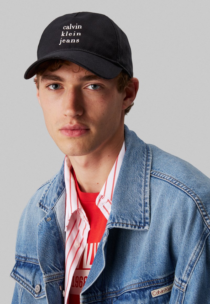 Juodas beisbolo kepurė su baltais "calvin klein jeans" tekstu, dėvima su džinsiniu švarku ant raudonos marškinėlių su baltais dryžiais, pabrėžianti sluoksniavimą.