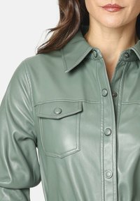 Mujer con una chaqueta de cuero verde con botones a presión y un bolsillo con solapa en la parte frontal, visible parcial entre los hombros y el pecho.
