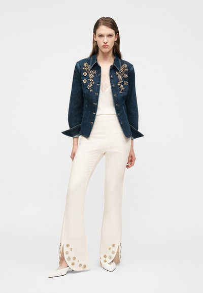 Γυναίκα που φορά μαύρο τζιν μπουφάν με χρυσές ανθισμένες διακοσμήσεις, λευκή τιραντέ μπλούζα και λευκό ανοιχτό παντελόνι με ανάλογες χρυσές ανθισμένες λεπτομέρειες.