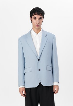Lyseblå blazer med strukturert design, enradet lukking, to frontlommer og skreddersydd passform, kombinert med en hvit skjorte.