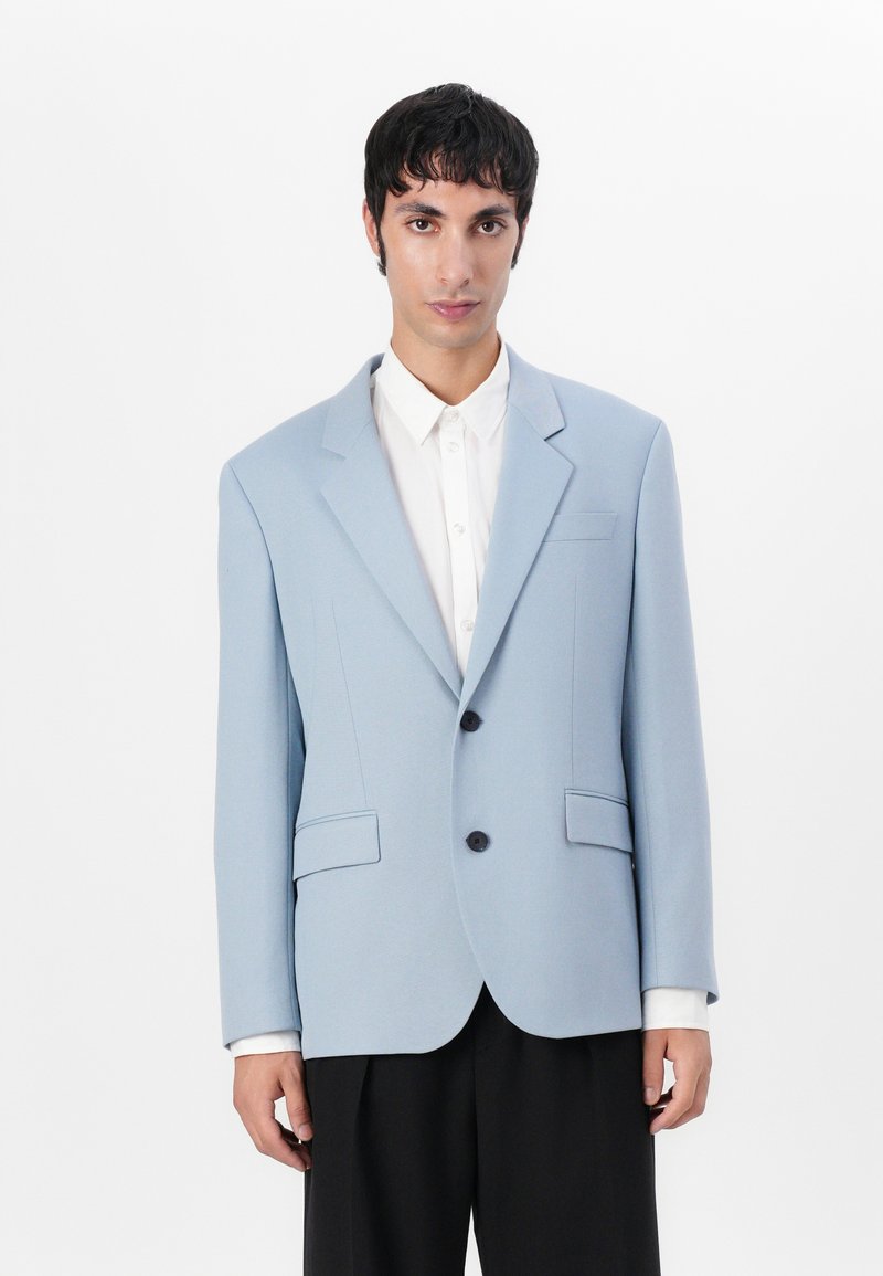 Helles blaues Blazer mit strukturiertem Design, einreihigem Verschluss, zwei Fronttaschen und taillierter Passform, kombiniert mit einem weißen Hemd.