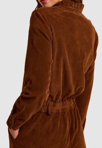 Brązowa kurtka z korduroyu z pionowym prążkowaniem, usztywnionym kołnierzem i elastycznym pasem. Posiada zapinane na guziki mankiety oraz boczne kieszenie.