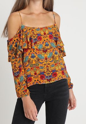 Femme portant un chemisier orange vif à épaules dénudées avec des motifs floraux multicolores et un pantalon noir sur un fond uni.
