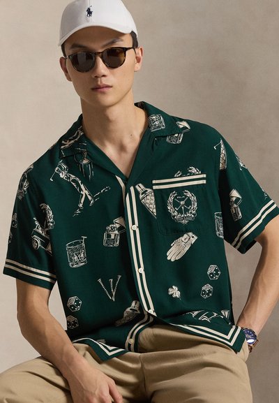 Młody mężczyzna ubrany w ciemnozieloną, wzorzystą koszulę z krótkim rękawem, beżowe spodnie, białą czapkę, okrągłe okulary przeciwsłoneczne i niebieski zegarek na nadgarstku, siedzący na tle jednolitego tła.