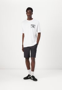 Hvid bomulds t-shirt med sort "Mickey" grafik, parret med mørke shorts og sorte sneakers med hvide detaljer. Ensfarvet baggrund.