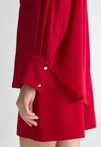 Blusa rossa con maniche lunghe e dettagli sui polsini. Il tessuto sembra liscio, con due bottoni dorati decorativi sui polsini. Design dell'orlo sovrapposto.