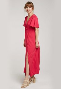 Robe fuchsia avec un décolleté en V profond, des manches flottantes et un détail de nœud à l'avant. Présente une fente à l'ourlet pour plus de mouvement.