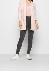 Blazer rosa claro con corte entallado sobre una blusa rosa con texto blanco, combinado con jeans skinny gris oscuro y zapatillas altas blancas.