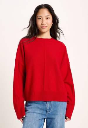 Young woman with long dark hair wearing a bright red sweater and blue jeans, standing against a plain light background.