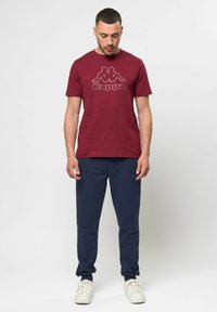 Burgundi Kappa póló, elöl logóval, tengerész kék jogger nadrággal és fehér sportcipővel párosítva, sima háttér előtt állva.