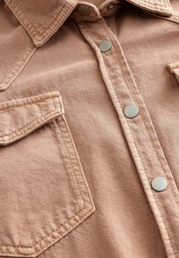 Cămașă din denim de culoare maro deschis, având guler, nasturi metalici argintii și două buzunare pe piept cu margini cusute. Textura este netedă și rezistentă.