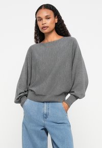 Suéter cinza de malha com um decote largo e mangas balão, combinado com jeans de denim azul claro. Textura suave e ajuste descontraído.