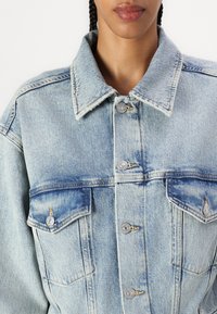 Lys blå denimjakke med klassisk krage, to brystlommer med klaffer og sølvfargede knapper. Utslitt tekstur over hele jakken.