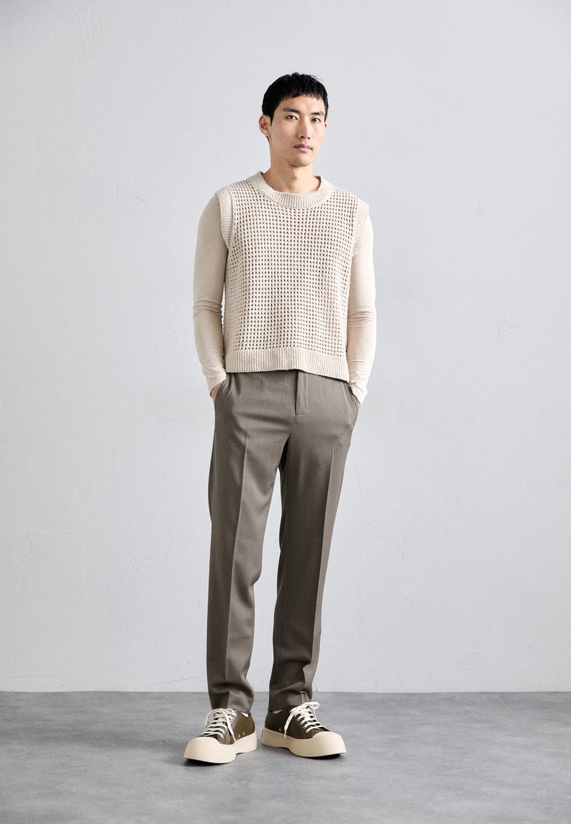 Homme se tenant debout les mains dans les poches, portant un haut sans manches en tricot beige sur une chemise à manches longues, un pantalon marron et des baskets à plateforme marron.