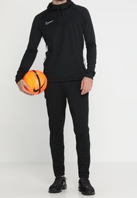 Schwarzer Nike Kapuzenpullover mit weißen Akzenten, kombiniert mit schwarzen Jogginghosen und schwarzen Sportschuhen. Hält einen orangefarbenen Fußball.