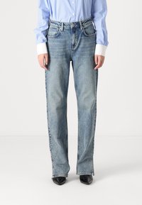 Person med ljusblå raka jeans, svarta spetsiga skor och en blårandig skjorta med vita manschetter, står framför en enkel bakgrund.