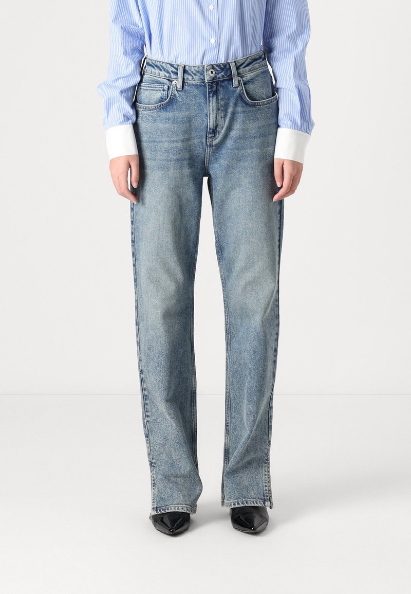 Person med ljusblå raka jeans, svarta spetsiga skor och en blårandig skjorta med vita manschetter, står framför en enkel bakgrund.