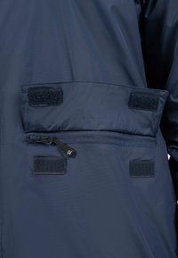 Marineblauwe waterdichte jasstof met een klittenband-flapzak en een ritszak met een zwarte ritstrekkertje en logo-detail.
