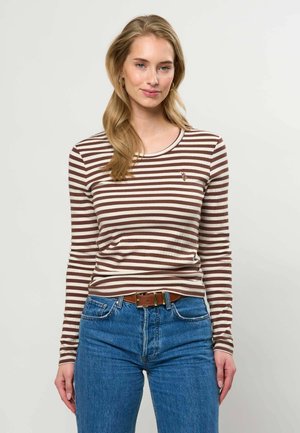 Kobieta o długich blond włosach, ubrana w brązowo-biały pasiasty longsleeve i niebieskie dżinsy z brązowym paskiem, stojąca na jednolitym tle.