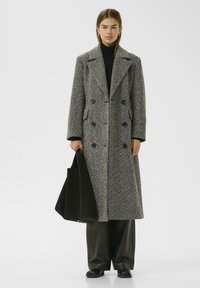 Abrigo de espiga gris con delantero de doble botonadura, grandes solapas con muescas y bolsillos laterales. Llevado sobre un jersey de cuello alto negro y pantalones negros.