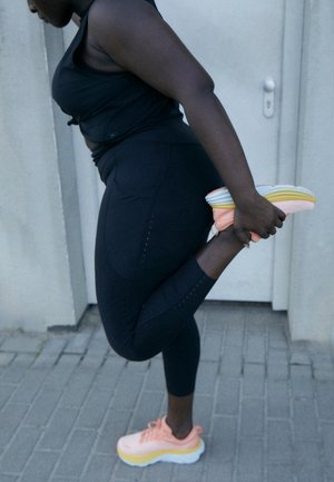 Black athletic leggings with dotted pattern, paired with light pink and yellow running shoes. Person stretches leg against a wall.