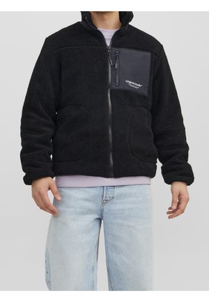 Man wearing black fleece jacket with zippered chest pocket and light blue jeans against plain background.