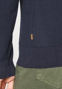 Mörkblå ribbade tröjornas fåll med liten orange "BOSS"-tagg, bärs med olivgröna strukturerade byxor och en synlig vänsterhand.