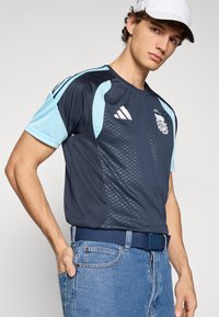 Adidas blue and navy sports jersey featuring short sleeves, mesh fabric, shoulder accents, and a crest logo on the front. Worn with denim jeans.