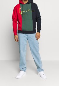 La felpa presenta un design a blocchi di colore con sezioni rosse, verdi e blu navy, una tasca a marsupio e un logo script giallo. Indossata con denim chiaro.