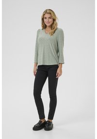 Un haut vert clair à manches longues avec un col en V et un motif à œillets, associé à un jean skinny noir et des mocassins à plateforme noirs.