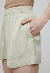 Lichtgroene shorts met een elastische tailleband, zijzakken en een gestructureerde stof. Gedeeltelijk bedekt door een hand.