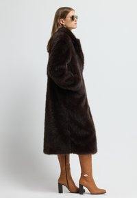 Abrigo de piel sintética marrón con un alto cuello y diseño en capas. Botas de cuero marrón hasta la rodilla con un acento dorado y tacones cuadrados negros. Gafas de sol verdes.