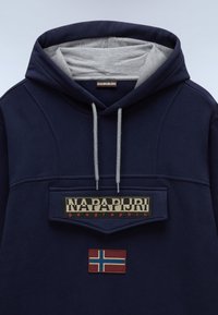 Sudadera de color azul marino con un interior forrado en gris, que cuenta con una gran etiqueta en el parche frontal y un emblema de la bandera noruega en rojo. Cuerdas ajustables en gris.
